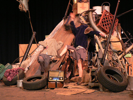 A chaotic pile of junk on stage with two people interacting energetically. The clutter includes tires, boxes, bicycles, and an old radio, evoking a sense of disorder.
