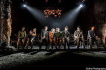 Seven people are seated in a dimly lit theatre, heads bowed under bright stage lights. The atmosphere conveys sombreness and introspection.