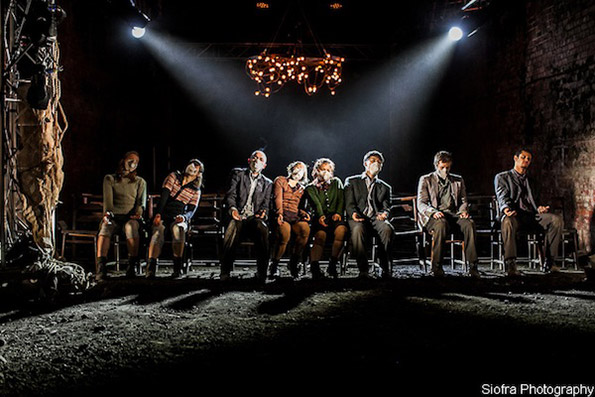 Seven people are seated in a dimly lit theatre, heads bowed under bright stage lights. The atmosphere conveys sombreness and introspection.