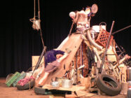 A performer climbs a slope made of junk, including tires, crates, and metal objects, against a black backdrop. The scene feels chaotic and industrious.