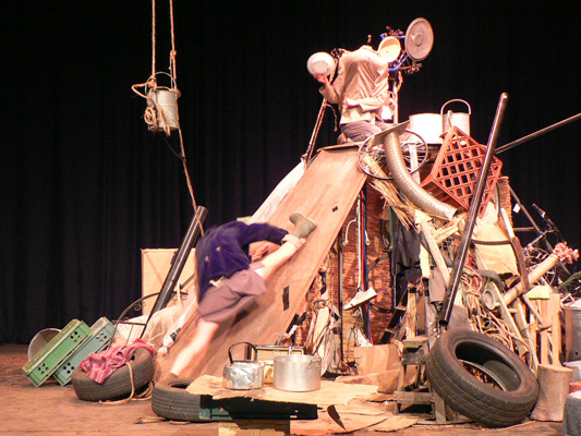 A performer climbs a slope made of junk, including tires, crates, and metal objects, against a black backdrop. The scene feels chaotic and industrious.