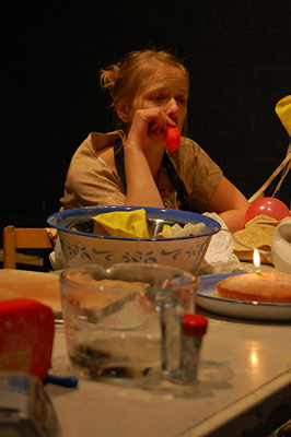 A woman sitting at a cluttered table, resting her head on her hand, looking contemplative. The table holds bread, a bowl, a glass, and a lit candle.