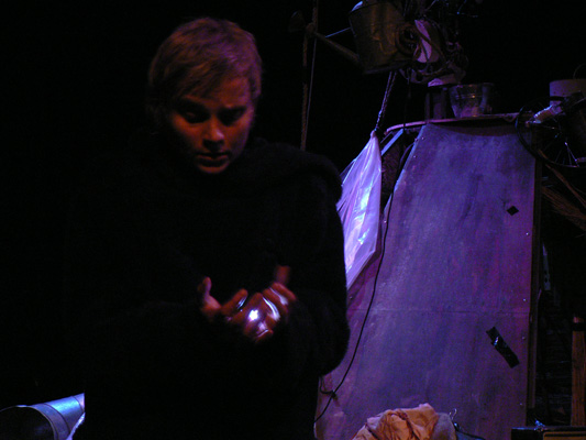 A person in dim lighting is centered, gazing at their glowing hands. A metallic structure with purple hues is behind them. The mood is mysterious.