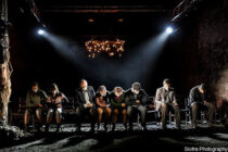 A group of people sits in a row on stage under dim lighting, casting shadows. The setting is dramatic and sombre, with a focus on emotion and intensity.