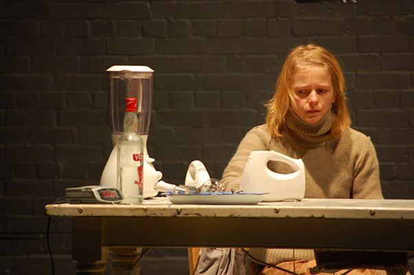 A woman with blond hair and a brown turtleneck sits at a table with a solemn expression. On the table are a blender, a vodka bottle, and a mixer.