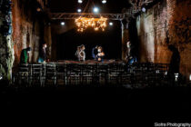 Dimly lit theater scene with a vintage chandelier hanging above. Seven people stand among scattered chairs on a rustic stage, creating a mysterious atmosphere.