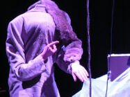 A person in a suit with a missing head illusion points at their wrist. A taxidermy animal is draped over their shoulders, in a dimly lit, surreal setting.