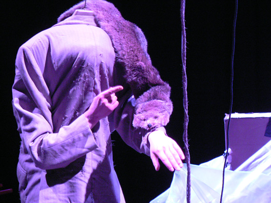 A person in a suit with a missing head illusion points at their wrist. A taxidermy animal is draped over their shoulders, in a dimly lit, surreal setting.