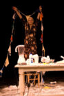 A woman joyfully holds colorful bunting on stage, surrounded by a blender and kitchen items on a table, set against a dark background.