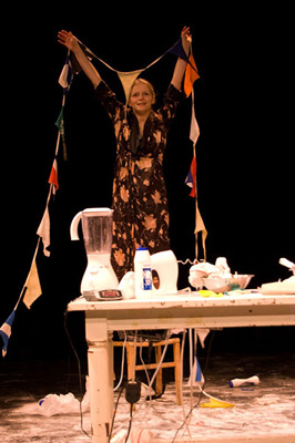 A woman joyfully holds colorful bunting on stage, surrounded by a blender and kitchen items on a table, set against a dark background.