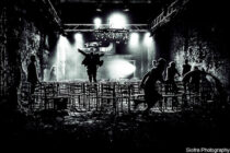 A black-and-white image of a dimly lit warehouse with people silhouetted against bright stage lights. One person is jumping onto a stack of chairs. The scene conveys energy and movement.