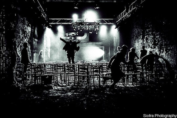 A black-and-white image of a dimly lit warehouse with people silhouetted against bright stage lights. One person is jumping onto a stack of chairs. The scene conveys energy and movement.