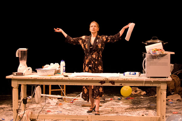 A woman stands on a cluttered stage in a messy kitchen setting, arms raised, conveying exasperation. Items like a blender and a microwave surround her.