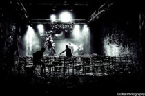 Dimly lit black and white theatre scene with scattered chairs and shadowy figures. Spotlight highlights activity, creating a mysterious, dramatic atmosphere.
