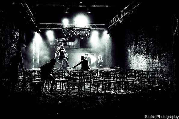 Dimly lit black and white theatre scene with scattered chairs and shadowy figures. Spotlight highlights activity, creating a mysterious, dramatic atmosphere.