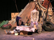 A chaotic stage set shows a pile of junk, including tires, cardboard, and a shopping cart. Two mannequins, headless and slumped, convey a sombre tone.