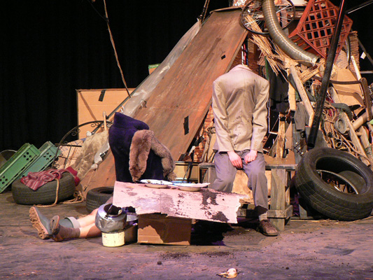 A chaotic stage set shows a pile of junk, including tires, cardboard, and a shopping cart. Two mannequins, headless and slumped, convey a sombre tone.