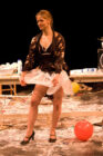 A woman in a playful pose stands on a messy stage with scattered flour, balloons, and cooking utensils. She wears a tutu, robe, and heels, exuding a humorous and lively vibe.