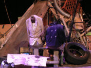 A dimly lit stage is cluttered with an eclectic mix of objects. Two headless mannequins, dressed in vintage coats, are positioned among old tires, metal pipes, a radio, and wooden scraps, creating a scene of organised chaos.