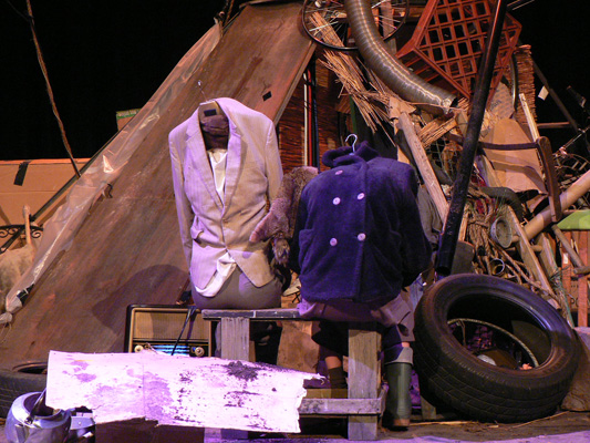 A dimly lit stage is cluttered with an eclectic mix of objects. Two headless mannequins, dressed in vintage coats, are positioned among old tires, metal pipes, a radio, and wooden scraps, creating a scene of organised chaos.