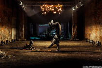 Break dancer performing a handstand in a dimly-lit industrial hall, illuminated by a chandelier. The mood is intense and dynamic, highlighting urban artistry.