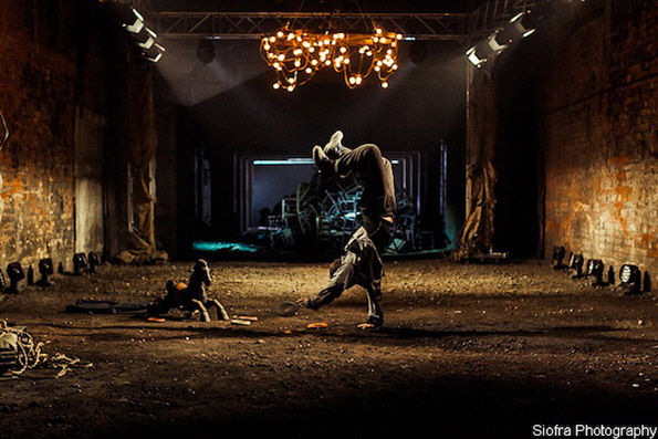 Break dancer performing a handstand in a dimly-lit industrial hall, illuminated by a chandelier. The mood is intense and dynamic, highlighting urban artistry.