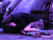 Person in dark clothing kneels and rests head on a tire amidst scattered debris. A pair of green boots lies nearby. Scene is dimly lit, evoking solitude.