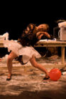 A performer in a dynamic pose leans against a table amidst scattered objects, wearing a white tutu and black top. A red balloon lies nearby on a cluttered floor.