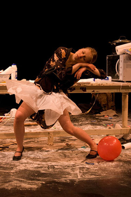 A performer in a dynamic pose leans against a table amidst scattered objects, wearing a white tutu and black top. A red balloon lies nearby on a cluttered floor.