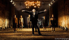 A person dramatically extends their arms in a dimly lit, industrial setting. Five others stand behind, illuminated by a warm chandelier overhead, conveying intensity.