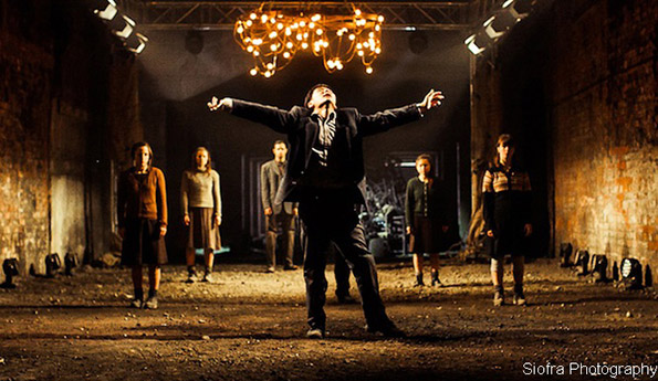 A person dramatically extends their arms in a dimly lit, industrial setting. Five others stand behind, illuminated by a warm chandelier overhead, conveying intensity.