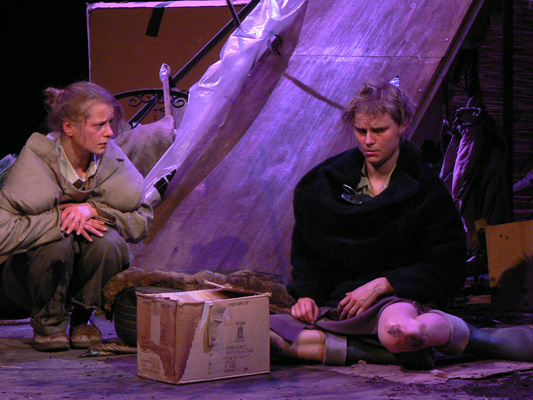 Two actors in a dimly lit theatrical scene convey tension. One person crouches, the other sits with a sombre expression. A cardboard box is nearby.