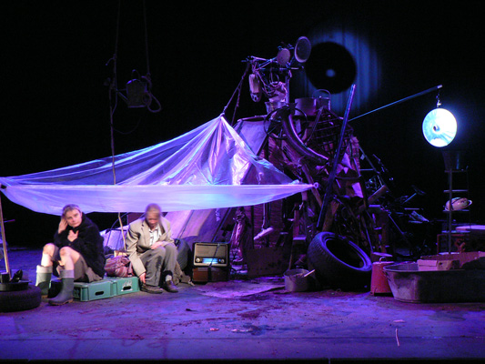 Two actors sit under a makeshift tent of translucent plastic amid a stage cluttered with tires and scrap. The scene is dimly lit, creating a sombre mood.