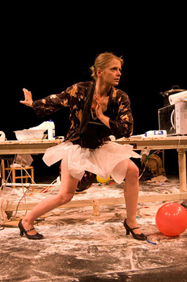 A performer in a dynamic pose on a cluttered stage, wearing a floral jacket and tutu. The scene is playful and chaotic, with scattered props and a red balloon.