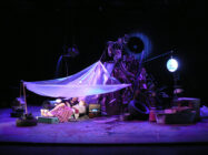 Dimly lit stage with a makeshift shelter of translucent fabric covers two people among scattered tires and debris, evoking a futuristic scrapyard vibe.