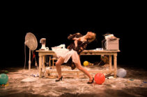 A woman in a dramatic pose leans back near a cluttered table with a fan, mixer, and scattered balloons. The scene conveys chaos and theatricality.