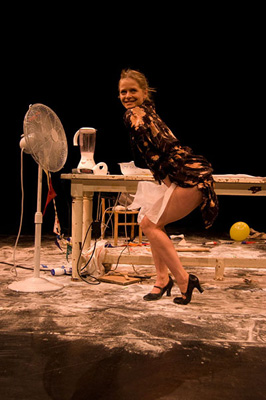 A woman in a floral top and white skirt bends slightly near a cluttered table with a fan, blender, and wires. She smiles widely, conveying a playful mood.