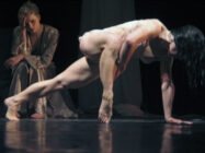A dancer performs a dynamic pose on stage, leaning on one arm with muscles tensed. Another person in a robe sits in the background, watching intently. The scene is dramatic and intense.