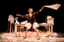 A performer strikes a dramatic pose in a cluttered kitchen setting. A fan blows on her, while she waves a cloth. The scene is chaotic yet lively.