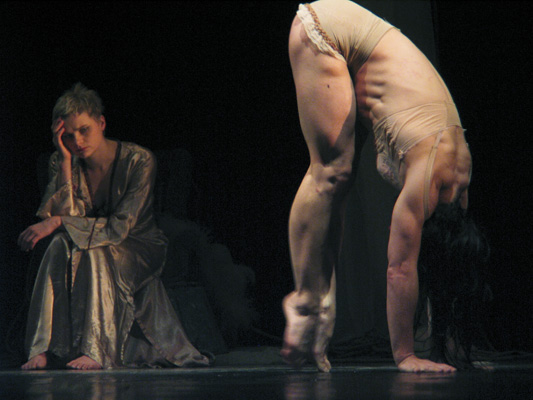 A contemplative seated performer in a flowing robe watches another in a revealing pose mid-dance, bending forward, showcasing fluidity and strength.