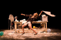 A performer dramatically leans back in front of a cluttered desk, surrounded by balloons, flour, and appliances. The scene conveys chaos and creativity.
