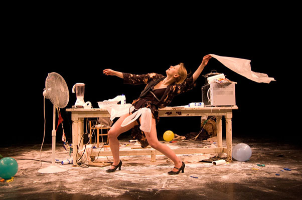 A performer dramatically leans back in front of a cluttered desk, surrounded by balloons, flour, and appliances. The scene conveys chaos and creativity.