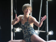 A performer with short hair and wearing a patterned dress poses expressively on stage, surrounded by bright stage lights, evoking a playful mood.