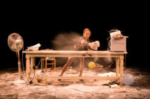 A woman energetically spreads flour on a cluttered table surrounded by scattered items, including a fan and balloons, creating a lively and chaotic scene.
