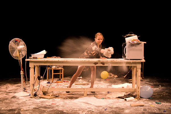 A woman energetically spreads flour on a cluttered table surrounded by scattered items, including a fan and balloons, creating a lively and chaotic scene.