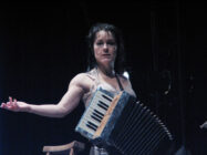 A woman stands on stage with an accordion, extending one arm expressively. The lighting is dramatic, creating a focused, intense atmosphere.