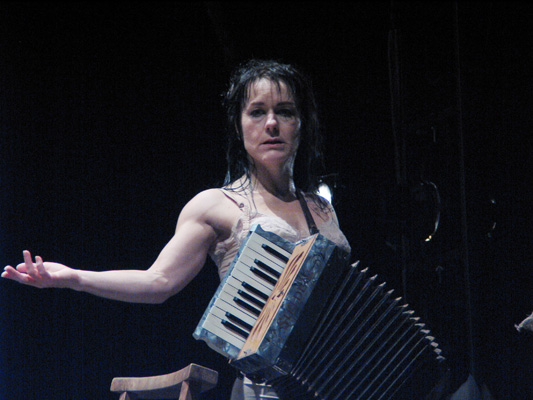 A woman stands on stage with an accordion, extending one arm expressively. The lighting is dramatic, creating a focused, intense atmosphere.