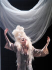 A performer in a dramatic pose wears an extravagant feathered headdress and gown. White drapery hangs above, adding elegance to the theatrical scene.