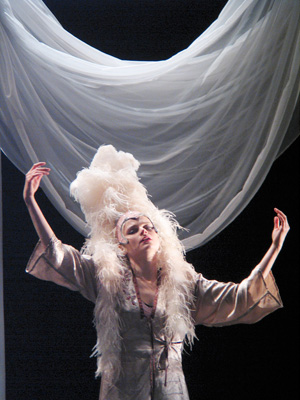 A performer in a dramatic pose wears an extravagant feathered headdress and gown. White drapery hangs above, adding elegance to the theatrical scene.
