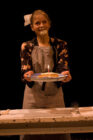 A woman with a bow in her hair, wearing an apron, playfully holds a plate with a lit candle. Her face is smeared with cream, conveying a light-hearted mood.